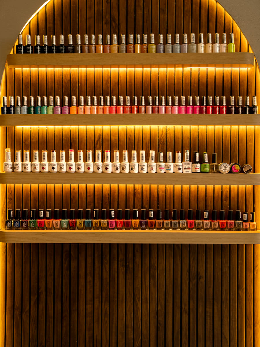 An arched wooden slat wall holding four backlit shelves of nail polish bottles in every colour.