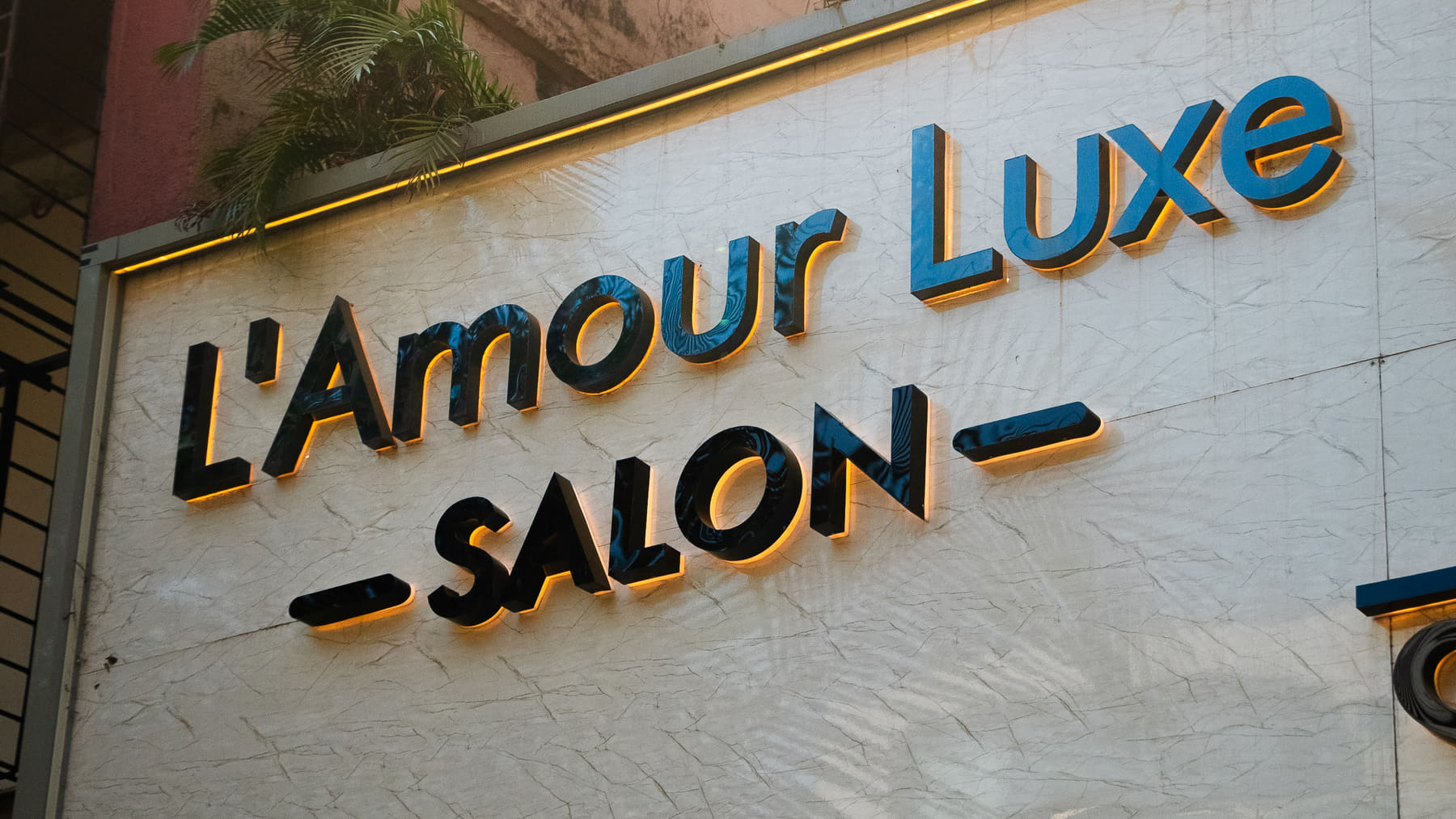 The salon's hand-painted shopfront sign reading L'Amour Luxe Salon, backlit in gold with late afternoon sun across it.