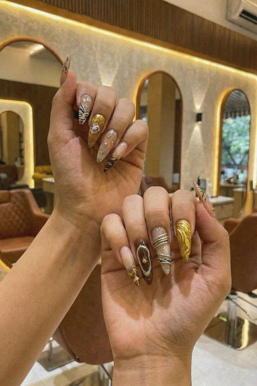 Two hands raised together showing long almond nail extensions in gold foil, chrome and brown agate art.