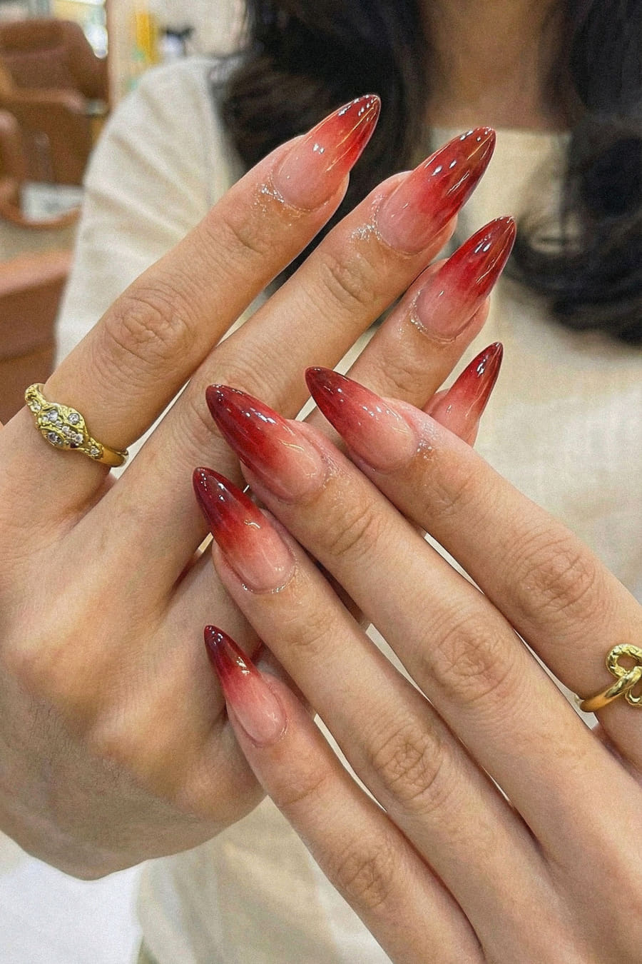 A close view of long almond nails in a deep red ombré fading to a bare tip, worn with gold rings.