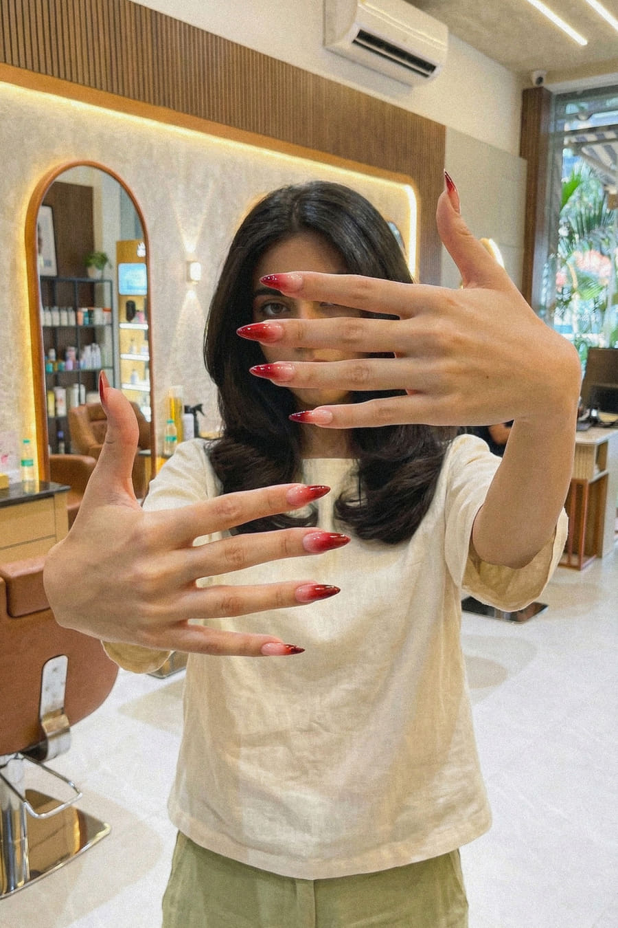 A client holding both hands up to the camera in the salon, showing a matching set of red ombré almond nails.