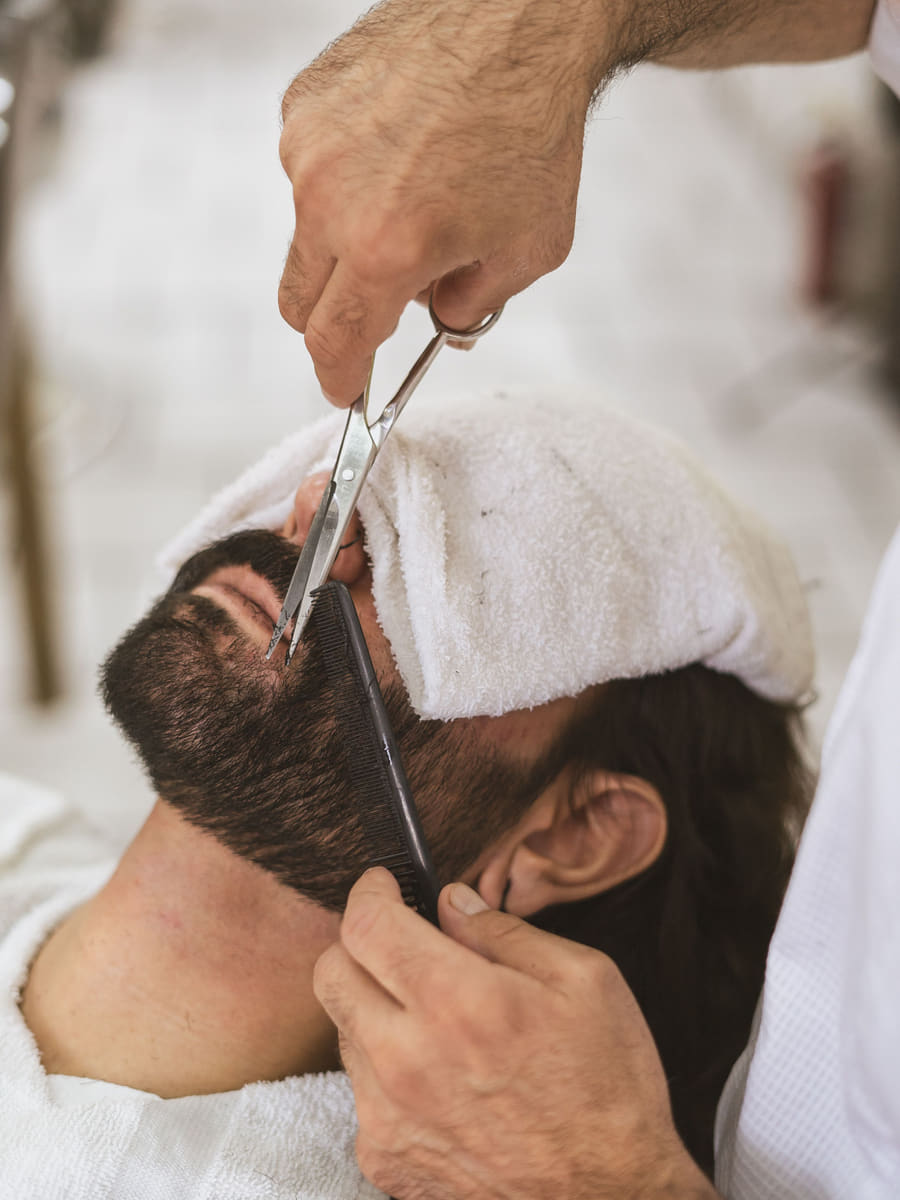 A barber trimming a client's beard with scissors over a comb, the client's face wrapped in a hot white towel.