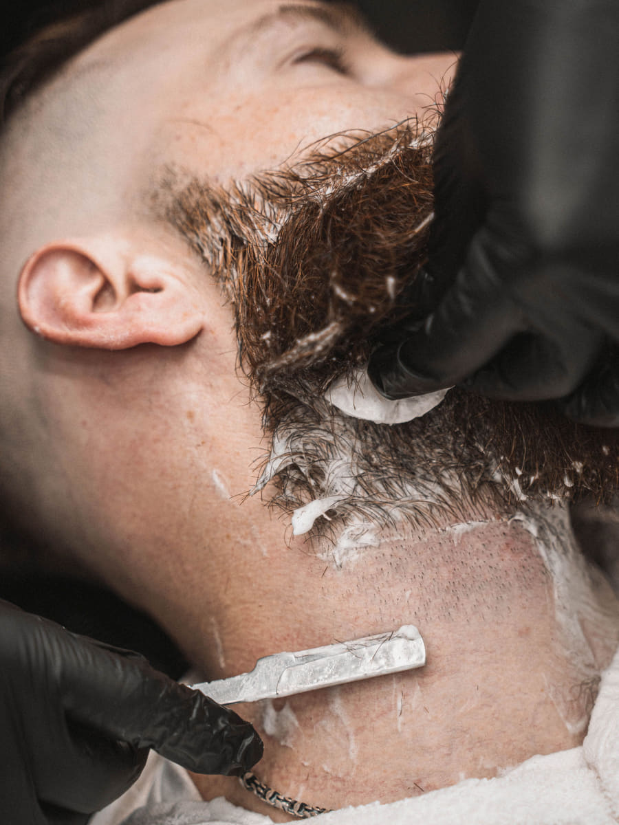 A close razor shave in progress along a client's neckline, the beard lathered and the barber working in black gloves.