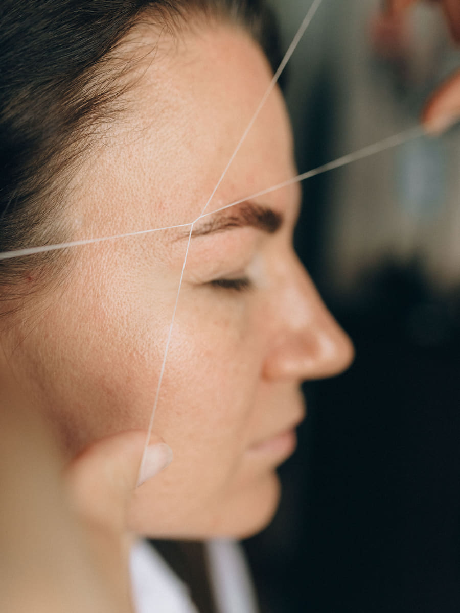 An eyebrow being shaped with cotton thread held taut across the brow, the client's eyes closed.
