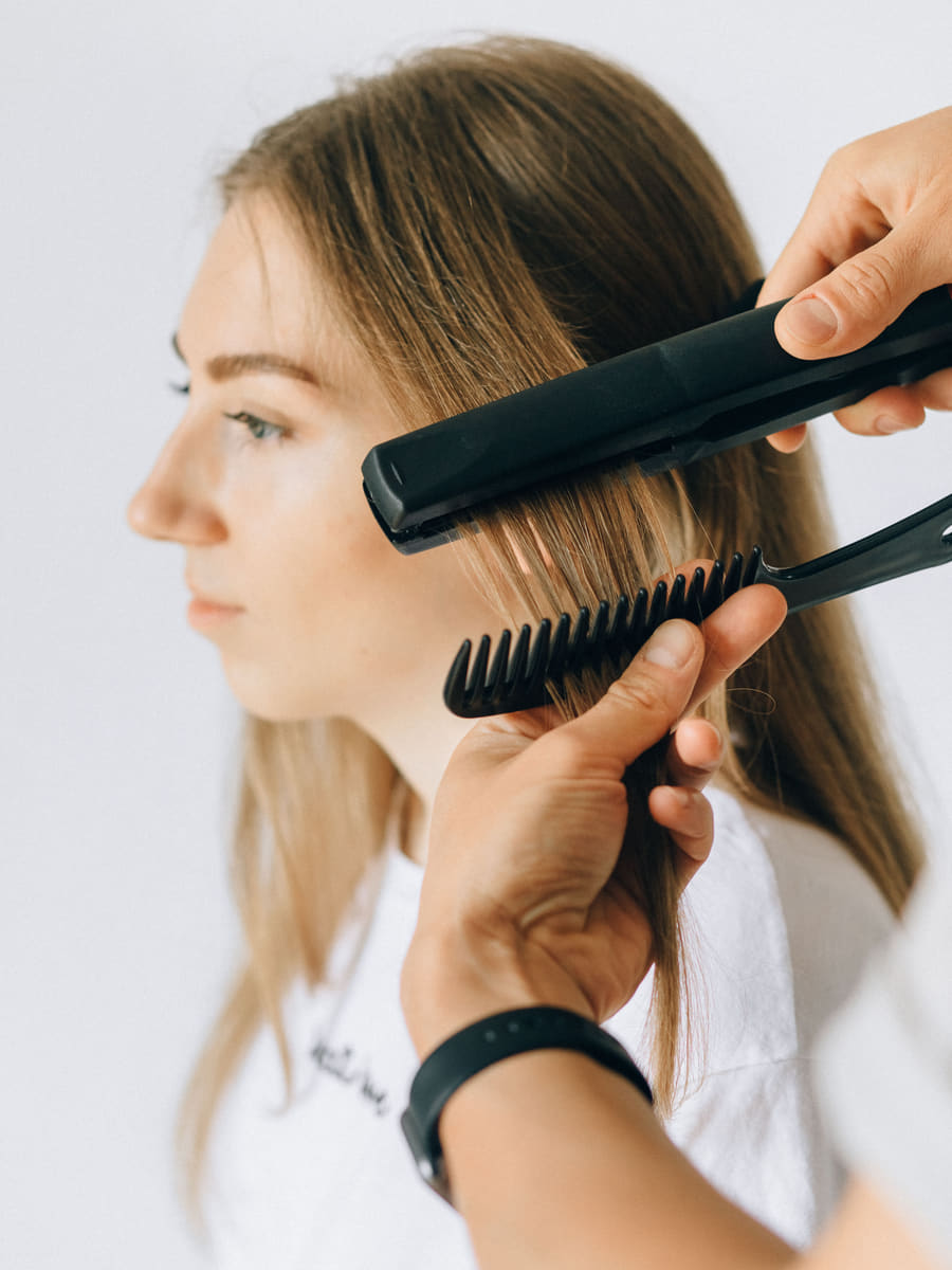 A stylist drawing a flat iron down a sectioned strand held on a comb, sealing a smoothing treatment.